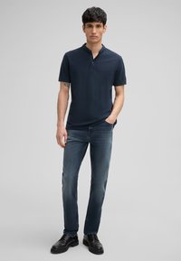 Navyblaues Baumwoll-Polohemd mit einem Henley-Kragen mit einem Knopf, kombiniert mit dunkelblauen Jeans und schwarzen Lederschuhen, vor einem neutralen Hintergrund stehend.