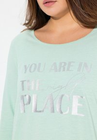 Mátově zelená dlouhá košile s dlouhými rukávy, na které je stříbrný metalický nápis "JSTE NA SPRÁVNÉM MÍSTĚ", s uvolněným výstřihem a jemnou texturou.