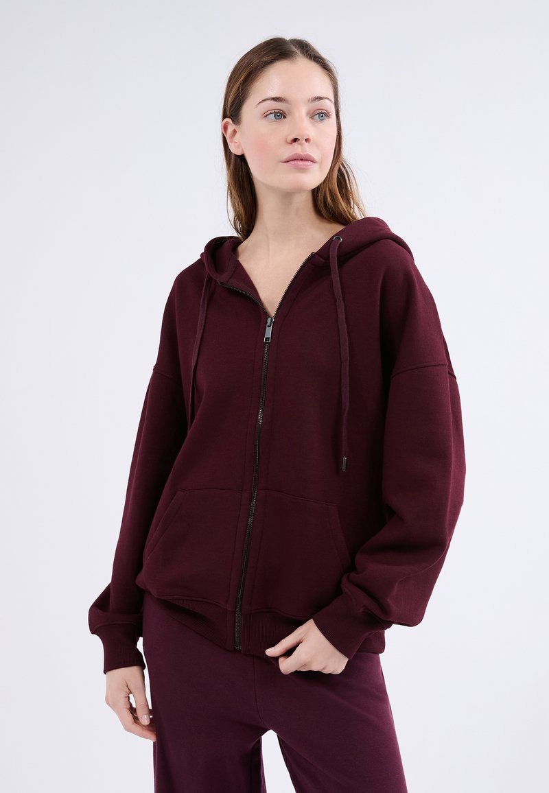 Felpa con zip bordeaux in un tessuto morbido e texture, con cappuccio, tasche frontali e vestibilità rilassata. Presenta coulisse e polsini a costine.