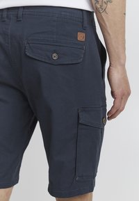 Shorts cargo bleu marine en tissu coton, dotés de poches latérales, d'accents en boutons et d'une poche arrière avec rabat, avec un détail de surpiqûre contrastante.