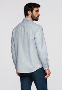 Homme aux cheveux foncés et à la barbe, portant une chemise à manches longues gris clair et un jean bleu foncé, de profil sur un fond uni.
