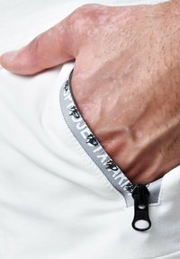 Bolsillo blanco con un detalle de cremallera gris, que cuenta con una lengüeta estructurada. Piel visible con vello fino, indicando un agarre firme en el bolsillo.