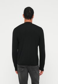 Vīrietis ar īsiem cirtainiem matiem, valkājot melnu džemperi ar garām piedurknēm un izbalējušas melnas džinsas, stāvot pret vienkāršu baltu fonu.