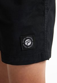 Pantalones cortos negros con una textura suave, que presentan una etiqueta rectangular negra con texto y logo blancos, cosida en el lado izquierdo.