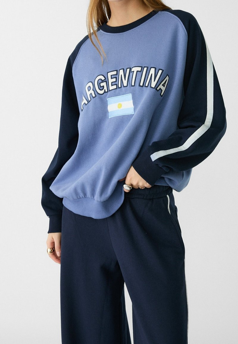 Personne portant un sweat-shirt bleu avec "ARGENTINE" et son drapeau sur le devant, associé à un pantalon bleu marine foncé avec des bandes blanches sur les côtés.