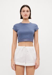 Světle modrá croptop košile s krátkými rukávy má texturovaný povrch a logo na lemu, kombinovaná s bílými texturovanými šortkami.