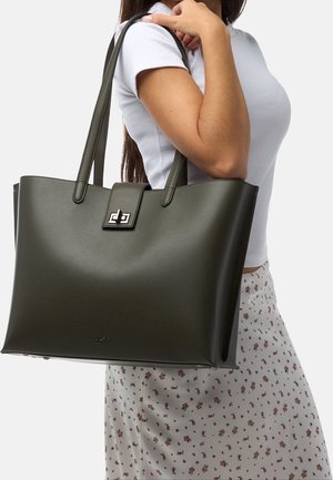 Olivengrøn shopper i tekstureret læder med sølvfarvet logo-lås og to lange håndtag, holdt af en person i en hvid skjorte.