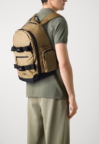 Mochila marrón con acentos negros, que cuenta con correas ajustables, múltiples bolsillos y un acabado texturizado. Se lleva en la espalda, con un diseño aerodinámico.