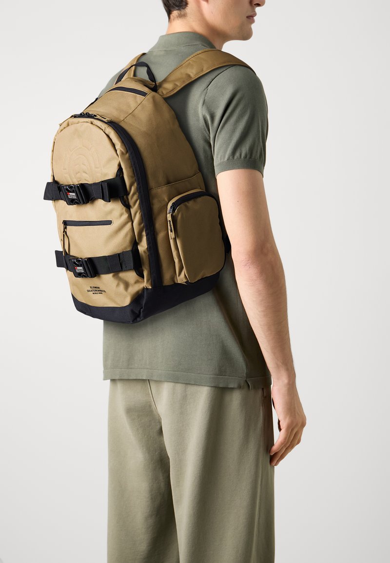 Mochila marrón con acentos negros, que cuenta con correas ajustables, múltiples bolsillos y un acabado texturizado. Se lleva en la espalda, con un diseño aerodinámico.