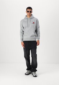 Grå hoodie med en forlommeslomme, der har en logo patch. Sammen med mørke jeans og grå sportssko. Modellen bærer solbriller.