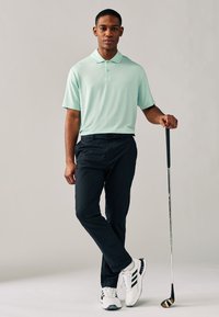 Mintgrön pikétröja, svarta byxor och vita golfsko med svarta detaljer. Modellen håller i en golfklubba. Mjukt tyg, modern passform.