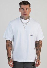 Camiseta de algodón blanca con mangas cortas, corte holgado, que presenta un pequeño logotipo negro en la zona izquierda del pecho. Textura suave, diseño minimalista.