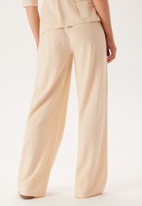 Lichtbeige wijdvallende broek gemaakt van zachte, textuurrijke stof. Heeft een ontspannen pasvorm met een hoge taille en een vloeiende silhouet.