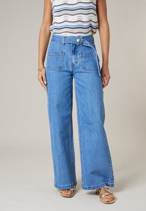Vrouw draagt blauwe jeans met wijde pijpen en hoge taille met grote voorzakken en een bijpassende riem, gecombineerd met een mouwloos gestreept gebreid topje.
