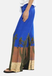 Niebieskie spodnie szerokie w nogawkach z motywem pustynnym, z zielonymi kaktusami i brązowymi detalami ziemi. Wykonane z lekkiego materiału, idealne na ciepłe dni.