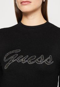 Schwarzer, gerippter Strickpullover mit einem 'Guess'-Logo aus silbernen und dunklen Steinen, mit klassischem Rundhalsausschnitt. Die Textur ist glatt und körperbetont.