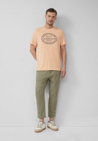 Licht perzik kleurig t-shirt met een grafische print in zwart, gecombineerd met olijfgroene broek en witte sneakers met tan accenten.