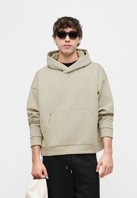 Νεαρός άνδρας που φοράει μπεζ φούτερ Puma και μαύρα γυαλιά ηλίου, κρατώντας ένα λευκό καπέλο στο αριστερό του χέρι, με απλό λευκό φόντο.