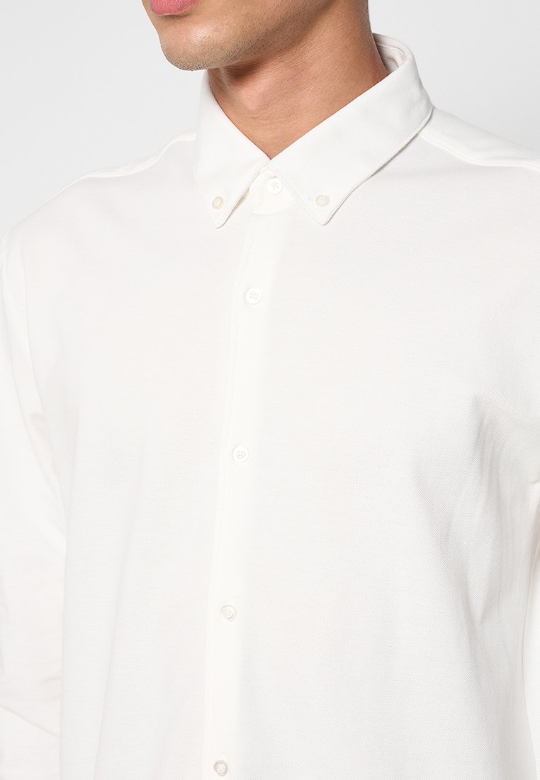 Biała koszula z guzikami z teksturowanej tkaniny, z wyraźnym kołnierzykiem i kilkoma guzikami na przodzie.