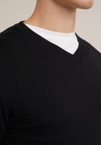 Musta neulottu V-kaula-aukollinen paita valkoisen paidan päällä, jossa on sileä pinta ja istuva muotoilu. Näkyvät saumat ja kaulusdetaljit.