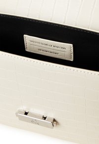 Biała torba nocna z teksturą krokodyla, z czarnym wnętrzem, zawierająca srebrny zamek oraz metkę z logo "United Colors of Benetton."