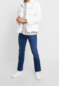 Valkoinen denim-takki, jossa on kaksi etutaskua, harmaa t-paita, tummansiniset farkut ja valkoiset lenkkarit. Rentoa muotoilua, jossa takin repaleiset helmat.