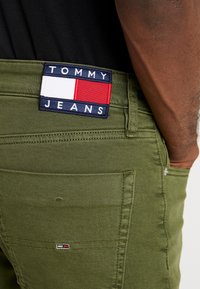 Olivgrön denimjeans med en tygpatch som har "TOMMY JEANS" i blått, vitt och rött. Inkluderar bakfickor och kontrasterande sömmar.