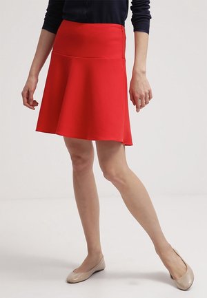 Femme portant une jupe rouge vif au genou, un haut bleu marine et des chaussures plates beige, debout devant un fond clair uni.