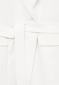 Vêtement blanc avec une ceinture en tissu nouée à la taille et un rabat rectangulaire de poche sur le côté droit.