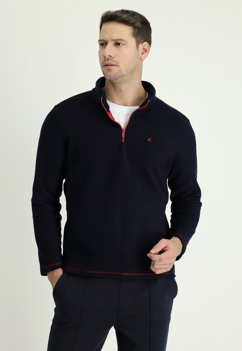 Marineblauwe pullover met een ritskraag, voorzien van rode accenten bij de rits en manchetten. Gemaakt van zachte stof met een effen structuur.