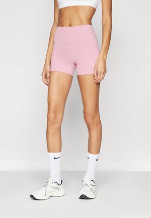 Kobieta ubrana w jasno-różowe sportowe spodenki z wysokim stanem, biały stanik sportowy, białe skarpetki Nike i białe sneakersy, stojąca na tle jednolitego jasnego tła.