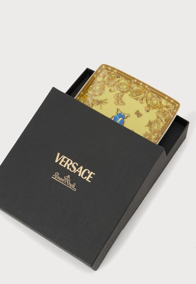 Μαύρο κουτί Versace με χρυσό λογότυπο, ημι-ανοιχτό, αποκαλύπτοντας ένα διακοσμητικό κίτρινο και χρυσό δίσκο με σχέδια έντομα και λουλουδιών στο εσωτερικό.