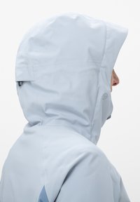 Veste imperméable bleu clair avec capuche, présentant une texture lisse, des détails de couture et un petit logo sur le côté. Vue de profil.