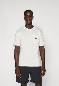 White cotton t-shirt featuring a chest pocket with a blue and white logo patch, paired with dark shorts and neutral background.