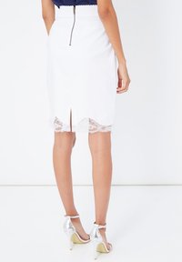 Falda lápiz blanca con cremallera trasera, dobladillo de encaje con forma de ondas y abertura en la parte posterior. Combinada con sandalias de tacón alto plateadas.