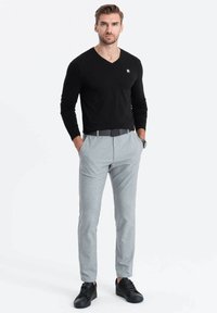 Maglietta nera a maniche lunghe con scollo a V, pantaloni grigi su misura e scarpe da ginnastica nere. La maglietta presenta un piccolo logo; i tessuti sono lisci e aderenti.