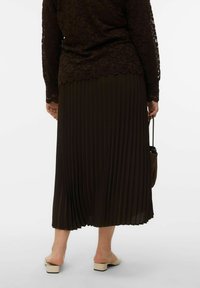 Jupe plissée marron foncé associée à un top en dentelle. La jupe présente une texture lisse et descend sous le genou, complétée par des sandales à talons carrés beiges.