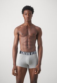 Pelēki kokvilnas bokseršortki ar melnu jostasvietu, uz kuras ir uzraksts "CALVIN KLEIN". Modelis stāv taisni, demonstrējot pieguļošu dizainu un gludu tekstūru.