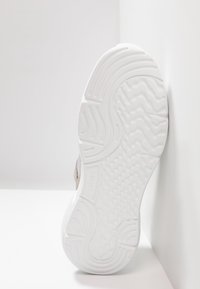 Biała podeszwa zewnętrzna trampka z falistym wzorem, z teksturowaną, wyściełaną powierzchnią i zakrzywionymi krawędziami, ustawiona pod kątem na jednolitym tle.