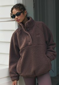 Pullover in pile marrone con colletto alto, zip frontale a metà e polsini elasticizzati. Texture morbida e branding discreto sul petto.