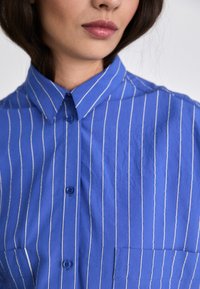 Camicia blu con righe verticali bianche, davanti con bottoni, colletto a punta e taschino sul petto. Tessuto morbido e leggermente stropicciato.