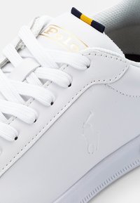 Vita lädersneakers med vita snörningar, med en gyllene "Polo"-logotyp och en subtil präglad hästlogotyp på sidan. Texturerad gummisula.