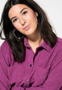 Camicia di velluto a coste viola con chiusura a bottoni, due tasche sul petto e colletto classico. Modella con capelli scuri e grandi orecchini a cerchio di argento.
