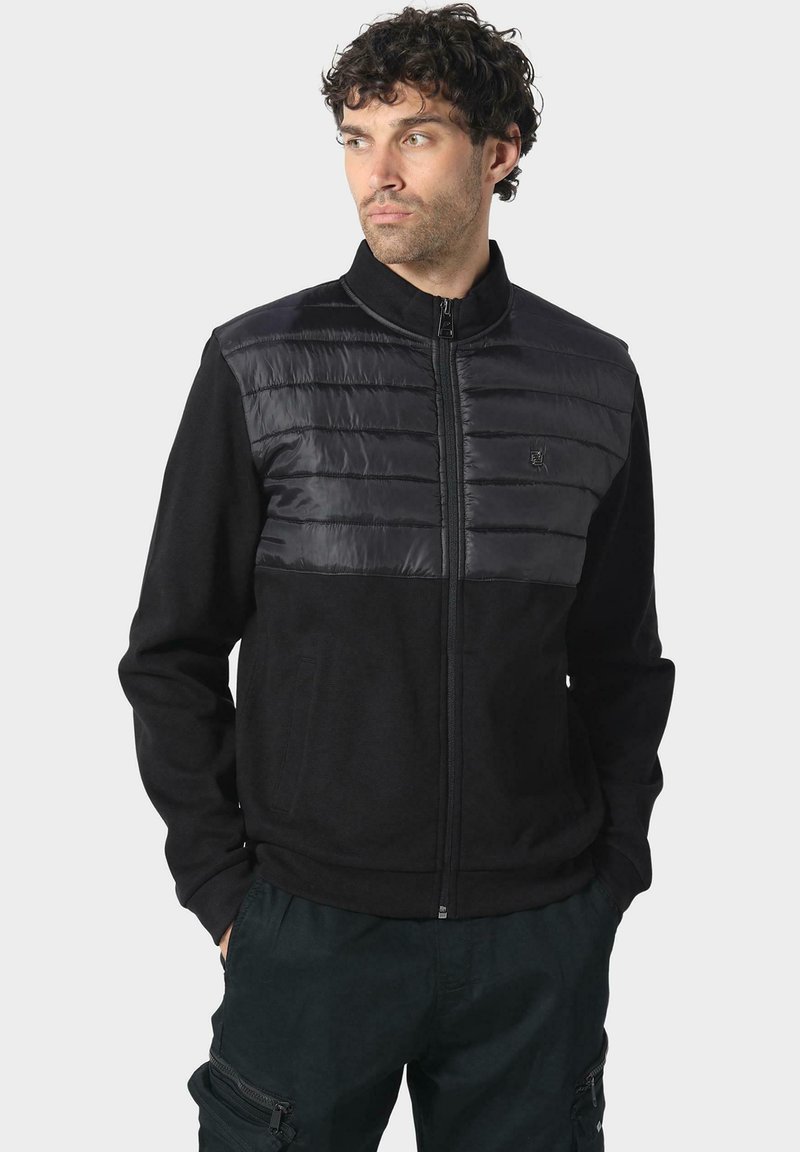 Chaqueta negra con la parte superior de nylon acolchada y la parte inferior de tela suave. Cuello alto, cremallera frontal y bolsillos laterales. Adecuada para uso casual.