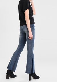 Žena v modrých džínách s rozparkem, černých vysokých kotníkových botách a černém tričku s krátkým rukávem stojí proti jednobarevnému světlému pozadí.