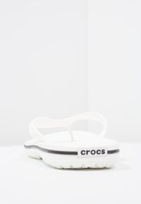 Valge kummist suss, millel on must triip, laia rihmaga disain ja soonikuga tallad. Brändi nimi "crocs" on nähtav külje peal.