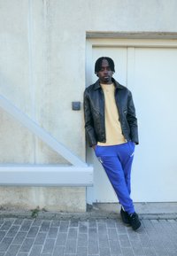 Czarna skórzana kurtka, żółta koszula, niebieskie spodnie dresowe z białymi paskami, stylizowane na czarnych trampkach. Stoi pod betonową ścianą.