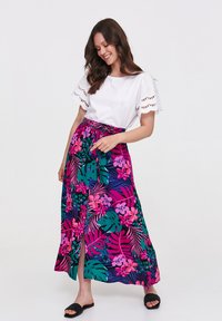 Maxi spódnica z intensywnym różowym i zielonym wzorem kwiatowym, wykonana z lekkiego materiału, w wysoki stan, związaną w pasie, z bocznym rozcięciem, w zestawie z białą bluzką.