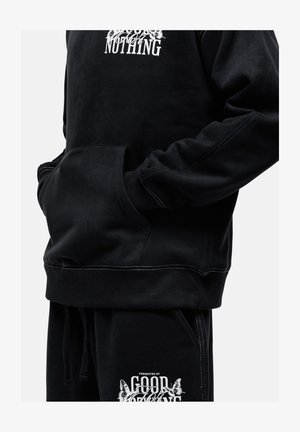 Felpa nera in pile con testo grafico bianco, dotata di tasca e polsini a coste. Abbinata a pantaloni neri con un logo bianco.
