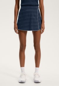 Navy modra pletena teniška krila z teksturiranimi črtami, kontrastnim belim obrobom in dekorativnim gumbom na pasu. V kombinaciji z belimi športnimi copati.